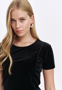 Czarna aksamitna koszulka z krótkimi rękawami i zaokrąglonym dekoltem. Posiada gładką teksturę i subtelny połysk, zestawiona z perłowymi kolczykami w kształcie kół.
