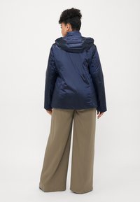 Kvinne som vender bort iført en marineblå hettegenserjakke og vide beige bukser foran en ensfarget hvit bakgrunn.