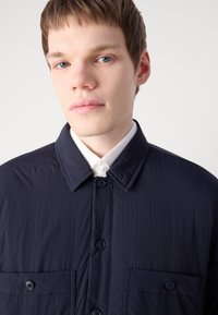 Camisa ligera de color navy con frente abotonado, dos bolsillos en el pecho, un cuello clásico y textura fina. Presenta botones azules.