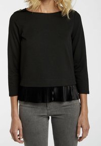 Svart långärmad topp med båthalsringning, med en plisserad satinkant nertill. Slät tyg med guldaccent vid axeln.