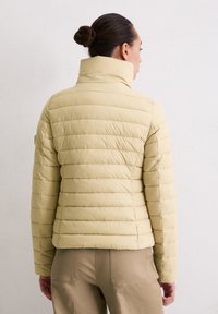 Beige, doorgestikte donsjas met hoge kraag en horizontale stiksels. Gladde stoftextuur. Achteraanzicht, gedragen met lichtgekleurde broek.