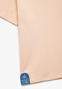 T-shirt z krótkim rękawem w kolorze brzoskwiniowym, z niebieską naszywką logo u dołu, przedstawiającą napis "RIO de JANEIRO" oraz wzór palmy.