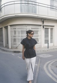 Crna košulja s kratkim rukavima s pojasom, u kombinaciji s bijelom teksturiranom suknjom s cvjetnim uzorkom, postavljena na urbanoj pozadini.