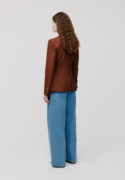 Bruine op maat gemaakte blazer met een gestructureerde afwerking, gecombineerd met lichtblauwe denim jeans met brede pijpen. Zichtbare knopen op de manchetten van de blazer.