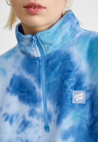 Blå tie-dye fleece-genser med halv-glidelås foran, myk tekstur og et firkantet logo-patch på brystet.