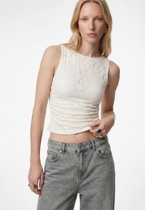 Kobieta w białym, bezrękawnym, koronkowym topie i szarych, wysokich dżinsach, z złotymi kolczykami i pierścionkami, pozująca na tle białego tła.