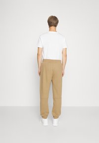 Beige Jogginghose mit einem Kordelzug am Bund, die sich an den Knöcheln verjüngt, kombiniert mit einem schlichten weißen T-Shirt und weißen Turnschuhen. Minimalistisches Design.