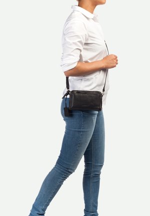 Bolso bandolera de cuero negro con acabado texturizado, compartimento con cremallera y correa desmontable, llevado en la cintura sobre jeans azules.