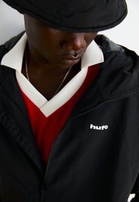 Black lightweight jacket with a zip, featuring a white logo. Underneath, a red v-neck shirt with a white collar and textured fabric.
