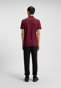 Bordeauxfarbendes Poloshirt mit kurzen Ärmeln und schwarzem Besatz, kombiniert mit schwarzen Hosen und dunklen Schuhen, von hinten betrachtet.