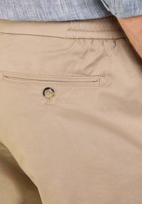 Pantaloni in cotone beige con vita elastica, dotati di una tasca posteriore chiusa da un bottone. Tessuto liscio con dettagli di cucitura sottili.