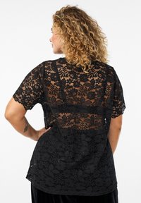 Top in pizzo nero con maniche corte, caratterizzato da motivi floreali e dettagli trasparenti sul retro. Tessuto morbido e vestibilità comoda.