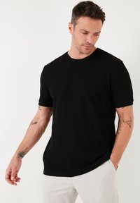 Čierny krátky rukáv tričko vyrobené z textúrovanej látky, s okrúhlym výstrihom a voľným strihom. Model stojí s rukou blízko vrecka.