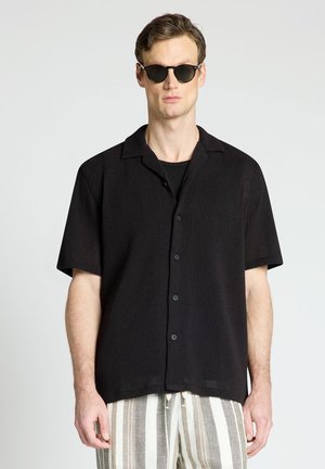 Uomo con camicia nera a maniche corte abbottonata, occhiali da sole scuri e pantaloni beige e bianchi a righe verticali, in piedi davanti a uno sfondo semplice.