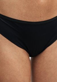 Gros plan d'une personne portant un bas de bikini noir avec une ceinture de marque indiquant "BOSS".