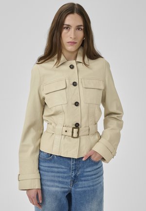 Pruunide pikkade juustega naine, kes kannab beeži vöökinnitusega jopet mustade nööpidega ja siniseid teksapükse, seisab ühevärvilise tausta ees.