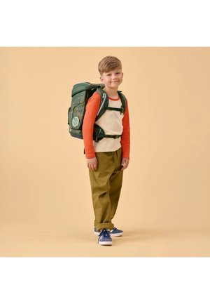 Junger Junge trägt ein orange-beiges Hemd, olivgrüne Hose, blaue Turnschuhe und einen großen grünen Rucksack, steht vor einem beigen Hintergrund.