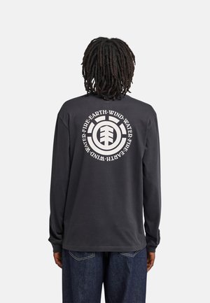 Persona con dreadlocks scuri indossa una maglia nera a maniche lunghe con un motivo circolare bianco e testo sulla terra, il vento, l'acqua e il fuoco sulla schiena.