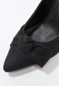 Chaussure à bout pointu en daim noir avec un nœud décoratif à l'avant, dotée d'une doublure intérieure lisse et d'une surface extérieure texturée.