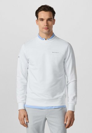 Man draagt een witte Hackett trui over een lichtblauw overhemd met kraag en lichtgrijze broek, staand tegen een effen lichte achtergrond.