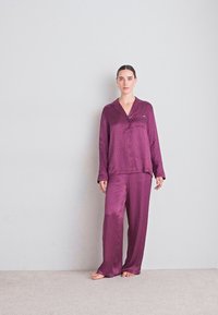 Satin-pajamasetti syvässä purppurassa, joka sisältää napitettavan yläosan, jossa on hienovarainen kuvio ja siihen sopivat löysät housut, pehmeä tekstuuri.