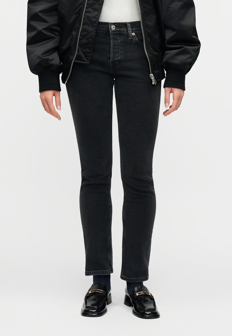 Άτομο που φοράει μαύρο τζιν στενής εφαρμογής, μαύρα μοκασίνια με χρυσή αγκράφα, μαύρο bomber μπουφάν και λευκό τοπ, στέκεται μπροστά σε λευκό φόντο.