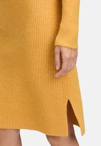 Hand, die entspannt an der Seite liegt, trägt ein senfgelbes, geripptes Strickkleid mit einem Schlitz an der Seite über dem Knie, helle Hauttönung, neutraler Hintergrund.