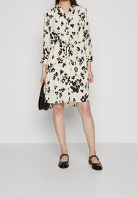 Kukkamekko vaaleassa kerma-värissä mustilla kuviolla, V-pääntiellä, pitkät hihat ja kapea vyötärö. Yhdistetty mustiin kiiltävän nahkaisiin Mary Jane -kenkiin.