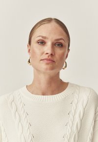 Biały sweter w sploty kablowe z chropowatymi wzorami i ściągaczem przy dekolcie; modelka z gładkim uczesaniem, złotymi kolczykami w kształcie kół oraz naturalnym makijażem.