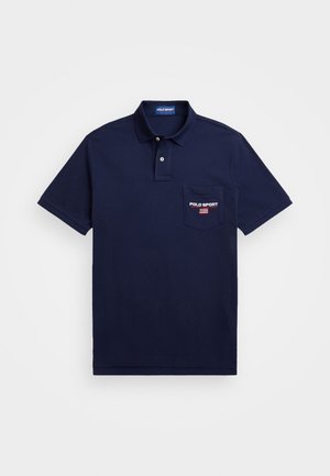 Mand iført navyblå Polo Sport polo med brystlomme og navy stribede bukser, hænderne i lommerne og armbåndsur på venstre håndled.