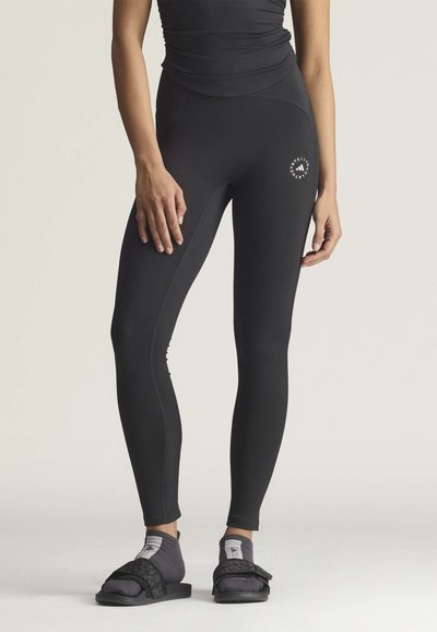 Kvinde iført sorte leggings med høj talje og logo på låret, kombineret med sorte sandaler og grå sokker, stående på en neutral baggrund.