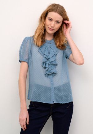 Lyseblå bluse i translucent materiale med korte ærmer, prikket mønster, flæser foran, knaplukning og rund halsudskæring. Bæres sammen med mørke jeans.