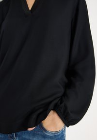 Czarna bluzka z dekoltem w kształcie litery V, długimi rękawami z elastycznymi mankietami i gładką fakturą tkaniny. Zestawiona z niebieskimi jeansami.