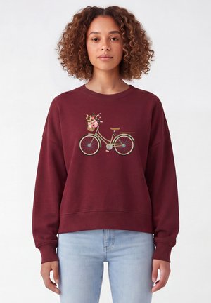 Jeune femme aux cheveux bouclés portant un sweat bordeaux avec un motif de bicyclette et panier de fleurs, associé à un jean bleu clair.