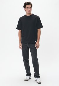Camiseta de algodón negra de mangas cortas, combinada con jeans de mezclilla gris oscuro y zapatillas blancas con detalles en gris, de pie contra un fondo blanco.