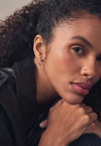 Nahaufnahme einer Person mit lockigem Haar, die eine schwarze Lederjacke und Creolen trägt. Die Haut hat eine glatte Textur, mit einem weichen Lippenfarbton.