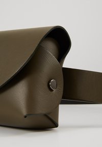 Gros plan d'une petite pochette en cuir vert olive avec un rabat et une fermeture à bouton rond argenté sur une sangle assortie.