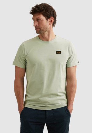 Homme portant un t-shirt vert clair à manches courtes avec un petit écusson logo noir et or sur la poitrine, les mains dans les poches, regardant sur le côté.