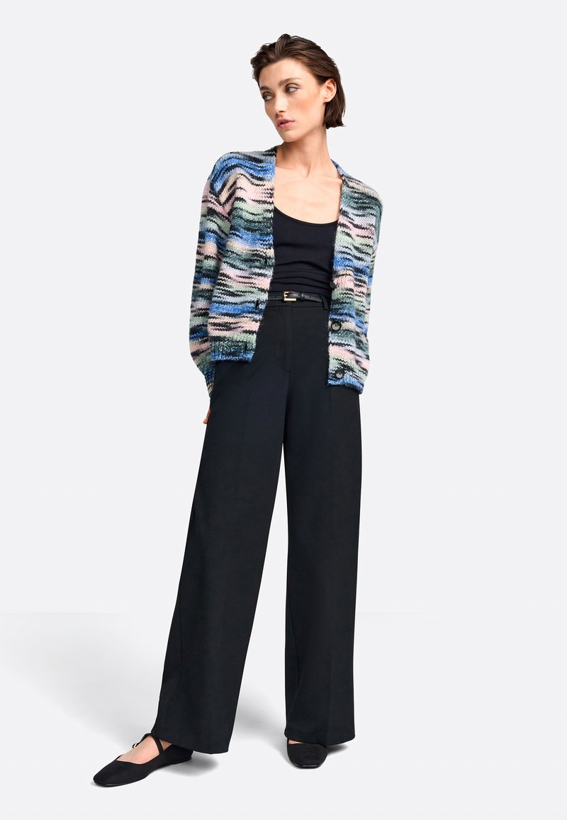 Multikleurige gebreide cardigan met golvende patronen, zwarte tanktop, hoge zwarte broek en zwarte ballerina's.