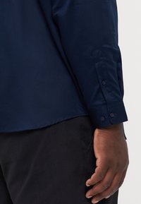 Camicia a maniche lunghe blu navy con una texture liscia e polsini con bottoni, abbinata a pantaloni neri, che mostra un primo piano della manica e della mano.