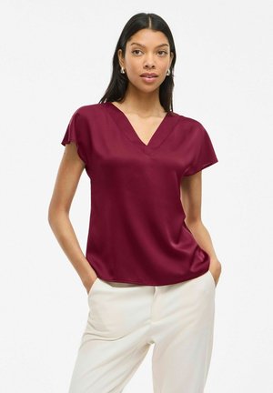 Burgundy V-neck blouse with short sleeves, made of smooth fabric, paired with cream trousers. Minimalist design, no visible patterns or accents.