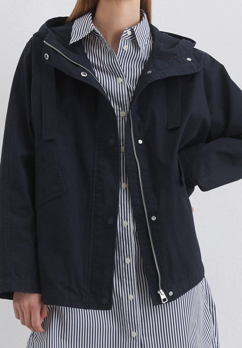 Femme portant une veste noire zippée par-dessus une robe chemise blanche à rayures verticales bleu marine.