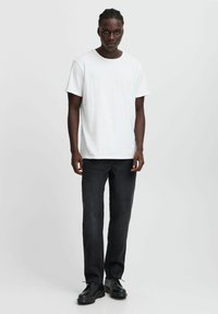 Camiseta blanca de manga corta combinada con jeans de mezclilla oscura y zapatos negros de cordones, exhibida contra un fondo claro y liso.