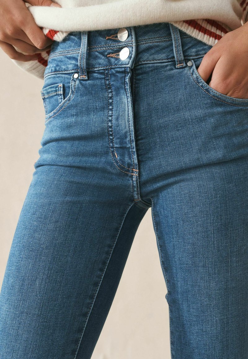 Gros plan sur une personne portant un jean taille haute bleu avec une fermeture à double bouton et les mains dans les poches avant.