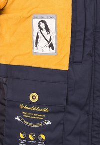 Veste bleu marine avec intérieur en polaire jaune, un patch montrant une femme avec une sangle d'épaule, et un texte imprimé décrivant les caractéristiques du tissu résistant à l'hiver.