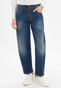 Donkerblauwe denim jeans met eenRelaxed-fit, voorzien van een knoopsluiting, vijf zakken en subtiele vervaagde delen op de dijen. Draagt met witte sneakers.
