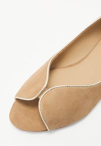 Beige zamšveida plakanas kurpes ar atvērtu pirkstu dizainu, izliektiem augšējiem malām un smalku metālisku apdari šuvēs. Gluda iekšējā oderes.