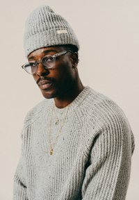 Šedý žebrovaný pletený svetr s kulatým výstřihem, doplněný šedou pletenou čepicí. Vrstvené zlaté náhrdelníky a brýle s průhledným rámem zvýrazňují vzhled.