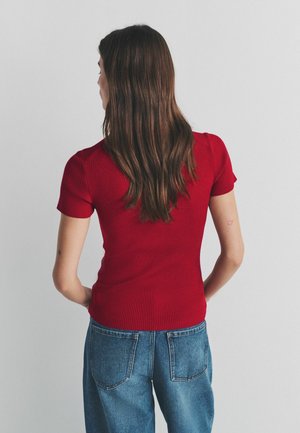 Woman with long brown hair wearing a fitted red short-sleeve top and blue jeans, standing with hands behind her back facing a plain light wall.