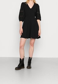 Vestido negro con escote en V, mangas cortas abullonadas con detalles de encaje, tejido ligero, forma en A; combinado con botas de combate negras con cordones.